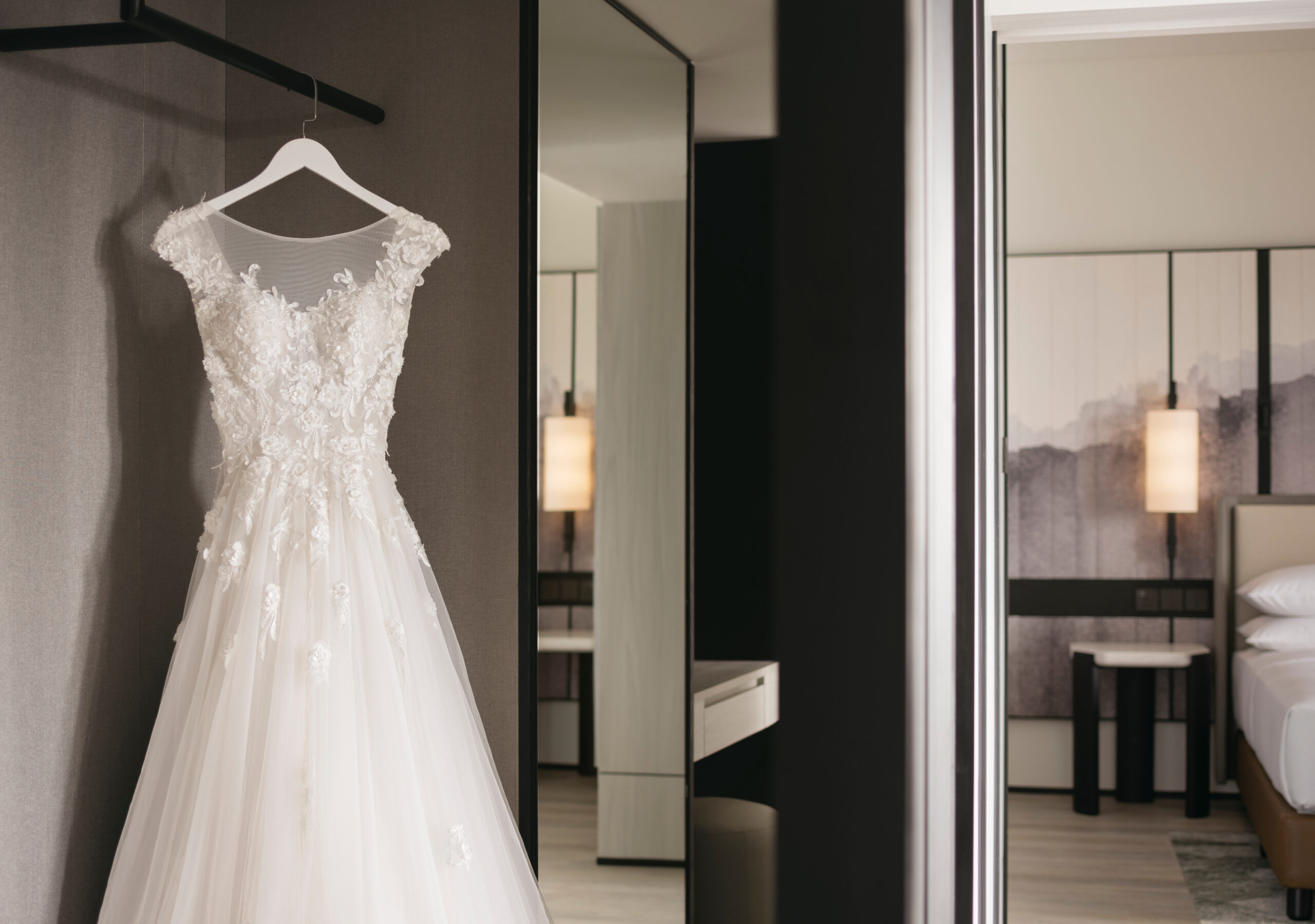 An ornate wedding dress hangs on a hanger against a neutral backdrop, with a stylish bedroom visible through a partially open door in the background.