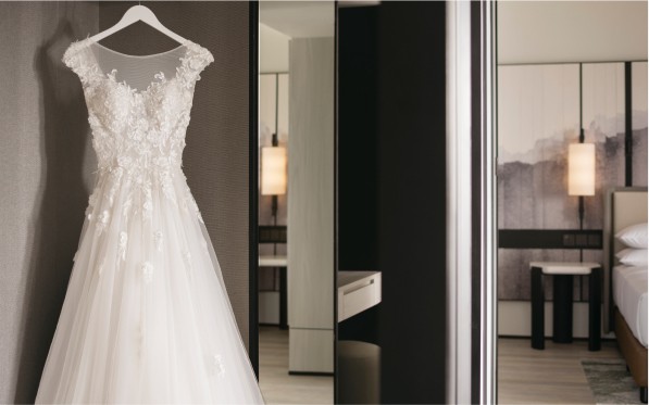 A white wedding dress with floral detail hangs in a modern bedroom; reflected in a full-length mirror adjacent to a neatly made bed and soft lighting.