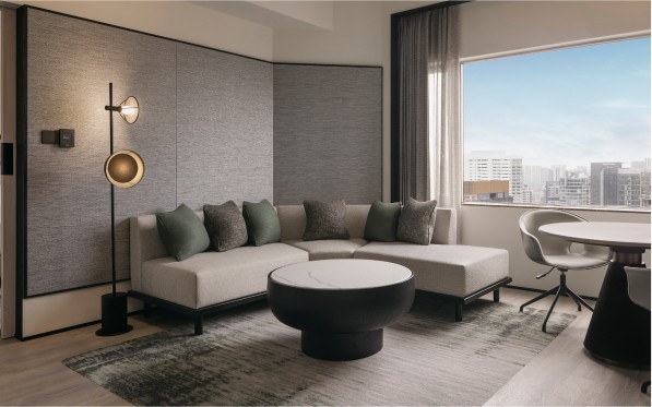 A modern living room features an L-shaped couch with cushions, a round coffee table, and a floor lamp, with a cityscape view through a large window.