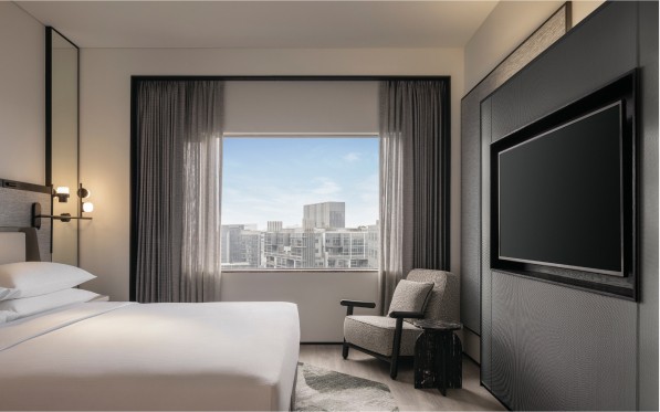 A modern hotel room with a bed, chair, side table, and TV, with large windows revealing a cityscape, showcasing a combination of comfort and urban living.