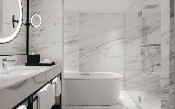 A modern bathroom with marble walls features a freestanding bathtub, a glass-enclosed shower, and a vanity with a round mirror, showcasing a clean and sleek design.