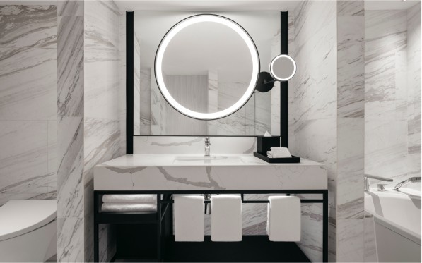 https://hiltonsingaporeorchardweddings.com/wp-content/uploads/2024/01/bridal6.jpg A modern bathroom vanity with a circular, illuminated mirror and a marble countertop, set against marble tiled walls. The space includes a faucet, towels, and a reflection of a toilet.