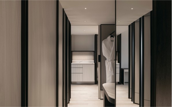 A modern hallway leads to a bedroom with a white-draped bed, reflecting a clean, minimalist interior design with dark and neutral tones.