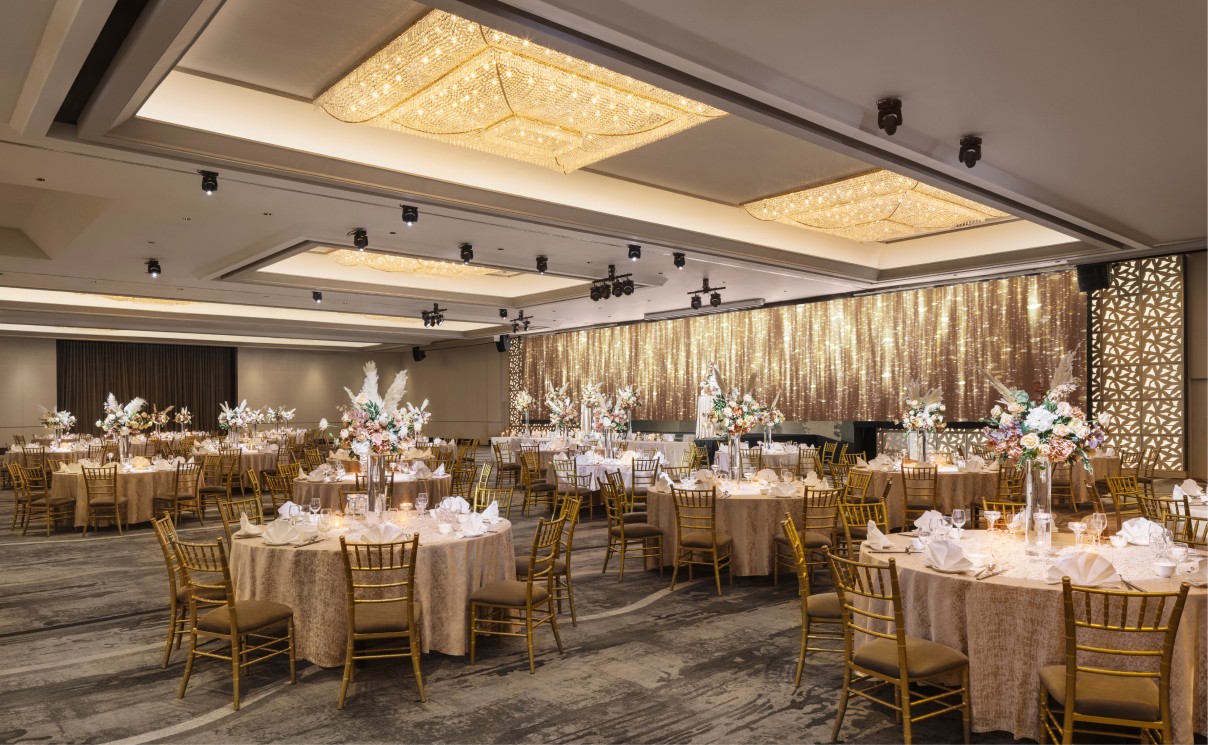 Elegantly set banquet hall with round tables, golden chairs, and floral centerpieces, under a warm chandelier illumination, awaiting guests for an event.