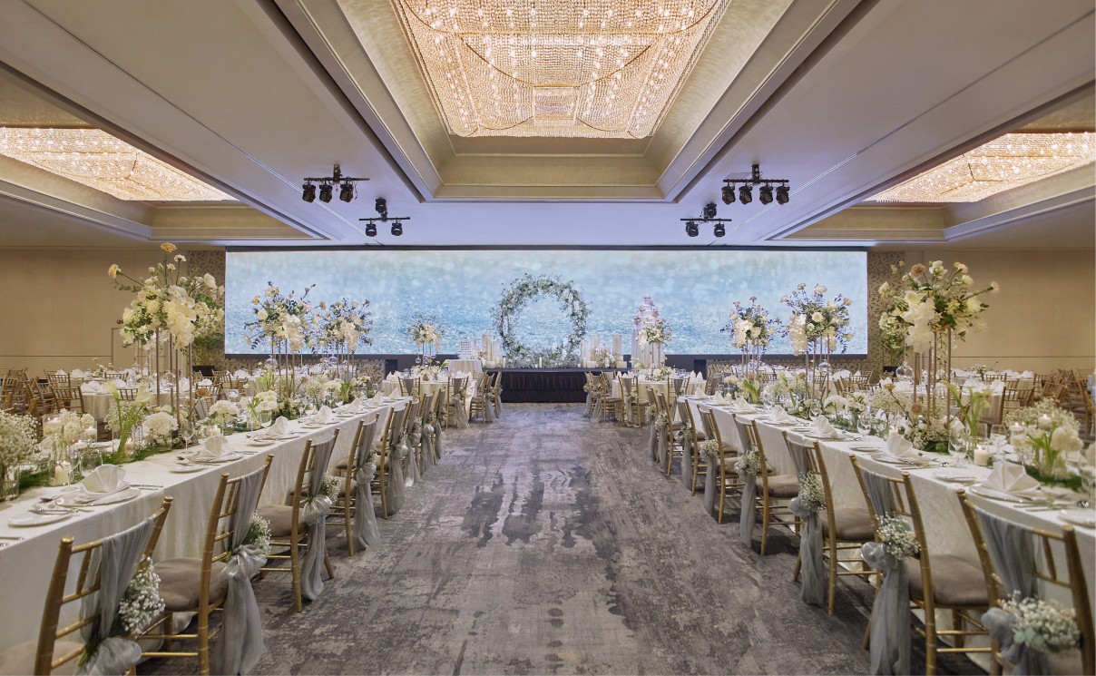Elegantly set tables in rows, adorned with white flowers and linens, ready for an event, inside a grand room with chandeliers and a floral arch backdrop.