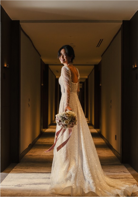 A woman in a long-sleeved, backless, glittering wedding gown smiles, holding a bouquet, in a warmly lit hallway.