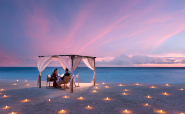 Two people dine under a canopy on a sandy beach at dusk, surrounded by lit candles, with a pink and blue sky over the ocean horizon.