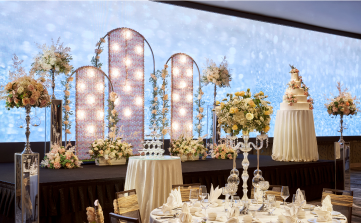 Elegantly set dining tables await guests in a room with floral decorations and a lighted backdrop, giving a warm, celebratory atmosphere, possibly for a wedding or formal event.