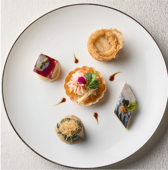 Five delicately plated gourmet appetizers are arranged on a white plate with a dark rim, presented on a light-colored tablecloth.