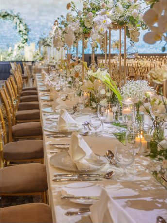 An elegant banquet table is set with fine dining cutlery, crystal glassware, and white floral arrangements, creating a luxurious atmosphere with a view of nature outside.