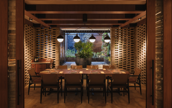A warmly lit dining area with a wooden table set for eight, surrounded by dark chairs. Overhanging pendant lights add ambiance to the space featuring brickwork and lush greenery outside.