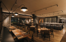 A stylish restaurant interior with tables set for dining, ambient lighting fixtures overhead, and a bar area in the background, all evoking a cozy, upscale atmosphere.