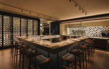 Modern restaurant interior with a large central bar, surrounded by stools, and walls lined with wine bottles; warm lighting creates a cozy ambiance. No text present.