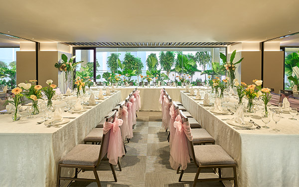 Elegant dining tables are set for an event, adorned with flowers and fine tableware, in a well-lit room with large windows showing lush greenery outside.