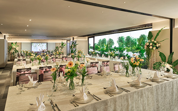 Elegantly set dining tables with white tablecloths, floral centerpieces, and place settings await guests in a bright, modern banquet hall with large windows overlooking greenery.