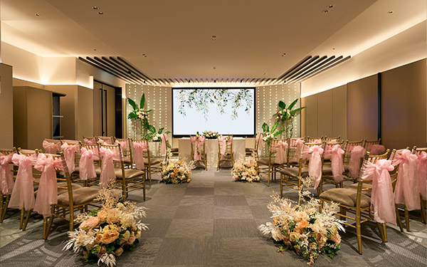 Rows of gold chairs with pink sashes flank an aisle adorned with floral arrangements, leading to a screen in a well-lit, elegant banquet hall.
