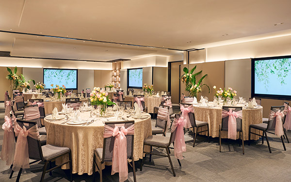 Elegantly set round dining tables with pink bows on chairs, arranged in a well-lit, modern banquet hall with large screens displaying leafy graphics.