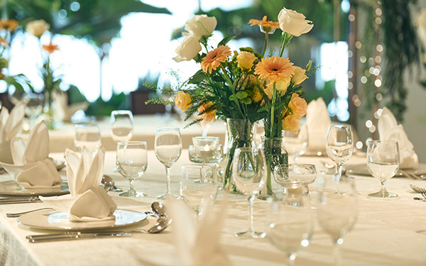 Elegant table setting with folded napkins, stemware, and a bouquet of white and peach flowers, poised for a formal event in a room with ambient lighting.