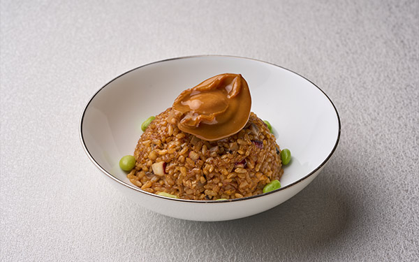 A mound of fried rice with green peas, topped with a dollop of brown sauce, served in a white bowl on a textured surface.