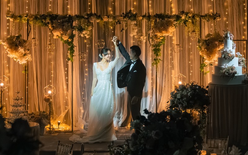 A couple dances amidst a romantic setting with fairy lights, candles, and floral arrangements, possibly at a wedding reception.